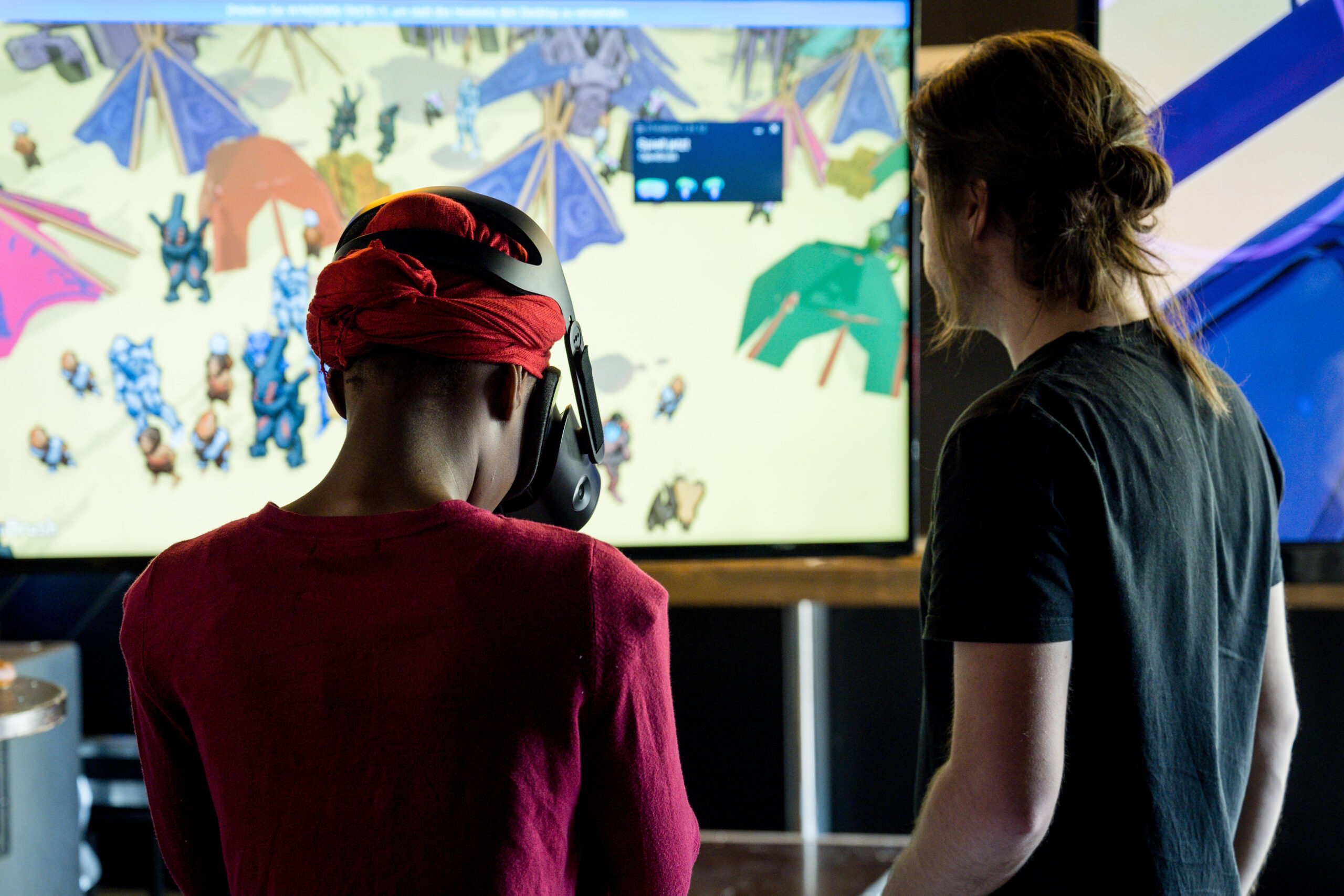 Zwei Personen vor einem Bildschirm. Die linke Person trägt eine VR Brille. Auf dem Bildschirm ist eine Landschaft und Figuren zu sehen.