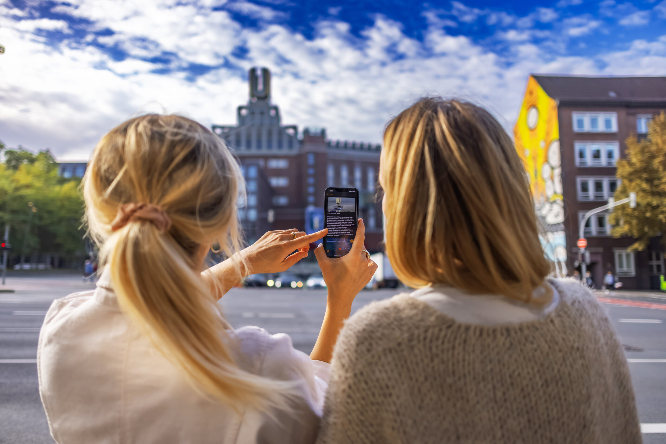 Zwei Frauen mit dem Rücken zur Kamera. Die eine hält ein Smartphone in der Hand auf der die Dortmunder Stadt-App zu sehen ist. Im Hintergrund sieht man das Dortmunder U